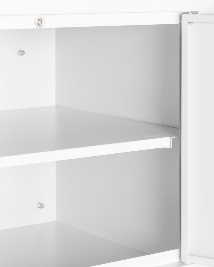 A close-up of an open, empty white wall cabinet with two shelves, viewed from the side. The cabinet door is partially ajar, revealing clean, smooth surfaces and metal mounting hardware securing the adjustable shelves in place. The background is plain and out of focus.