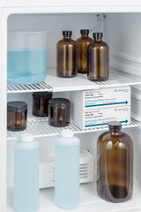 Small pharmaceutical compact refrigerator with brown glass bottles, clear jars with black lids, two white boxes labeled Influenza Virus Vaccine, and three clear bottles with blue liquid. Items are arranged on white wire shelves inside the medical refrigerator, which features a temperature alarm for added safety.