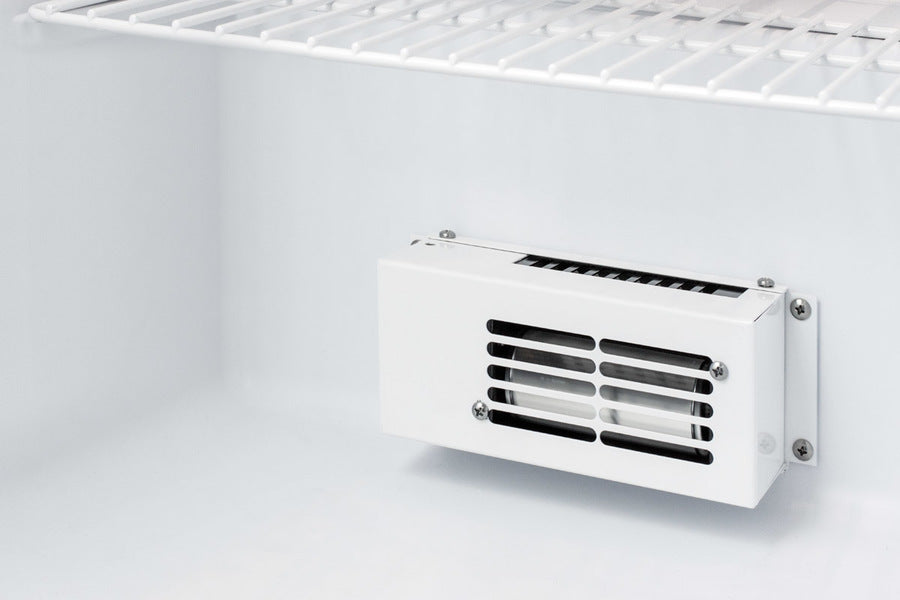 Close-up of a white, metal vent cover with horizontal slats, mounted on the back interior wall of a pharmaceutical compact refrigerator. Above is a white wire shelf. The scene is brightly lit, showing clean and empty fridge space.