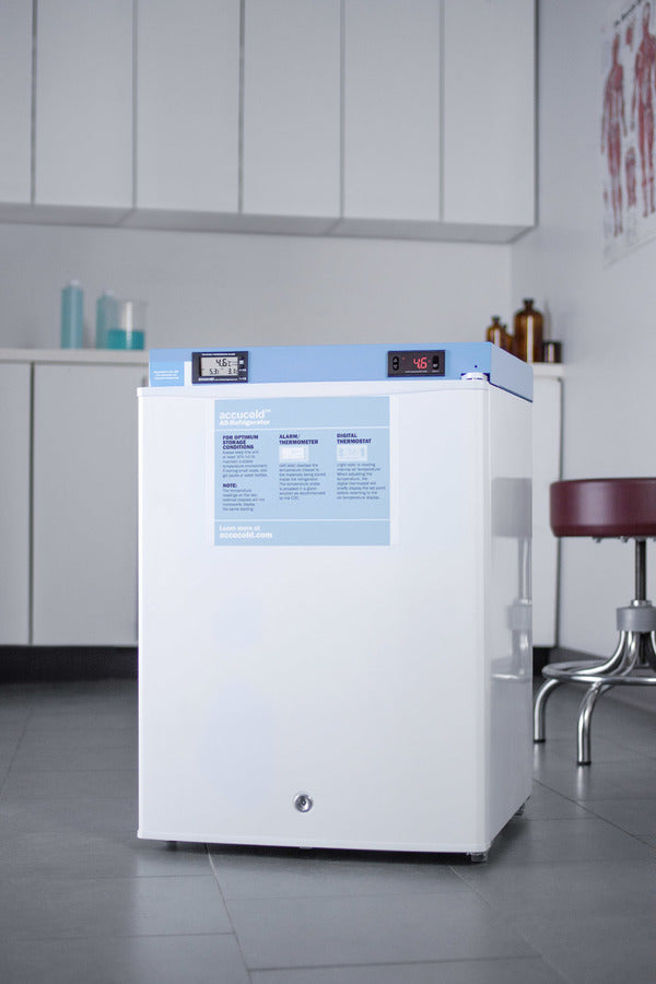 A pharmaceutical compact refrigerator with a blue top panel and digital display stands in a clinic room; this medical-grade refrigerator features a temperature alarm for added safety. Cabinets, jars, and a red stool are visible, along with anatomical posters on the wall in the background.