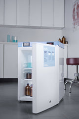 A small pharmaceutical compact refrigerator with an open door reveals clear and brown bottles inside. The fridge is situated in a clean, modern medical or laboratory room with cabinets, a stool, and medical posters visible in the background.