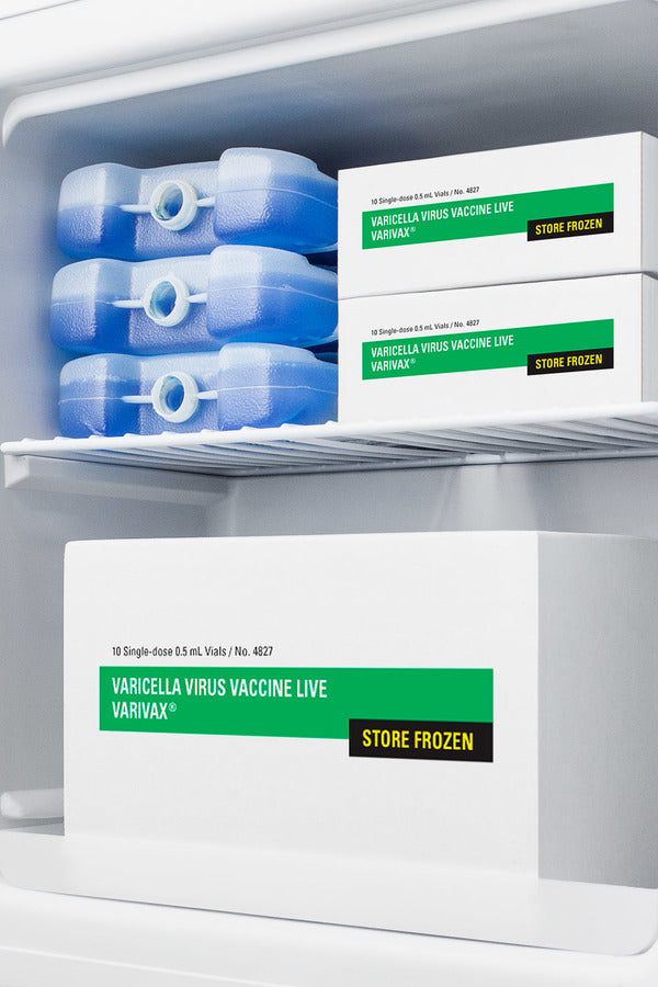 Inside an Accucold medical freezer, three blue ice packs are on the top shelf, next to two white boxes labeled Varicella Virus Vaccine Live VARIVAX Store Frozen. A larger box with the same label is on the bottom, against a white interior background. This compact all-freezer features NIST calibrated temperature monitoring for reliable vaccine storage.