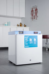 A white laboratory incubator with a blue control panel stands in a clean, modern lab alongside an Accucold medical freezer. Cabinets, jars, stools, and an anatomical poster are visible in the background. The floor is gray tile, and the room is brightly lit.