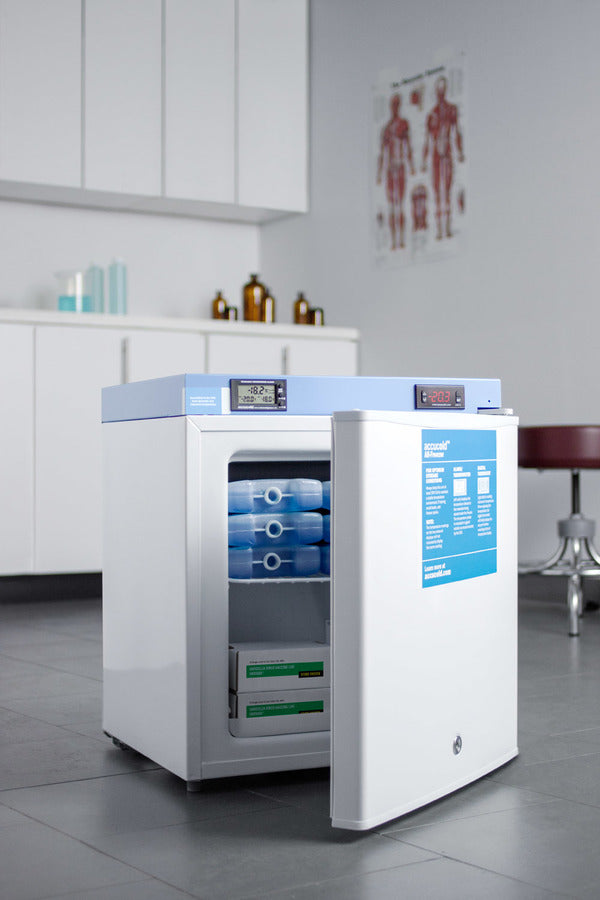 A medical-grade refrigerator stands open in a clean clinic room, revealing blue-packaged medical supplies and boxes inside. Cabinets, bottles, and a human anatomy poster are seen in the background. The gray tile floor complements the sterile environment, which also features an Accucold medical freezer—NIST calibrated to ensure precise temperature control for sensitive items.