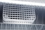 Close-up of a white, grid-like vent cover—possibly part of a commercial upright freezer or stainless steel freezer—secured with two screws and set against a reflective metallic surface. Bright white light from above creates reflections and lines on the metal, highlighting the sleek, utilitarian design typical of reach-in freezers.