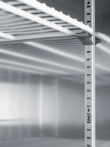 Close-up of an empty, metallic shelving unit inside a commercial refrigerator, with adjustable vertical supports marked with numbers. The photo is in grayscale, emphasizing the industrial and minimalist appearance of the shelves and the reflective surface behind them.