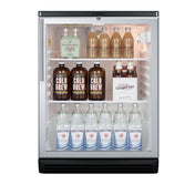 A small commercial glass door refrigerator with three shelves holds bottles of cold brew coffee, ginger ale, sparkling water, and other beverages, neatly organized in rows. The door is closed and the fridge is well-lit, showing the drink labels clearly.