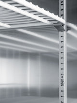 Close-up of a metal shelving unit corner, showing a numbered vertical support post with adjustable shelf slots. The parallel white shelves cast shadows on the metallic background, reminiscent of those found inside a commercial refrigerator. The environment is clean and minimalistic, suggesting industrial or storage use.