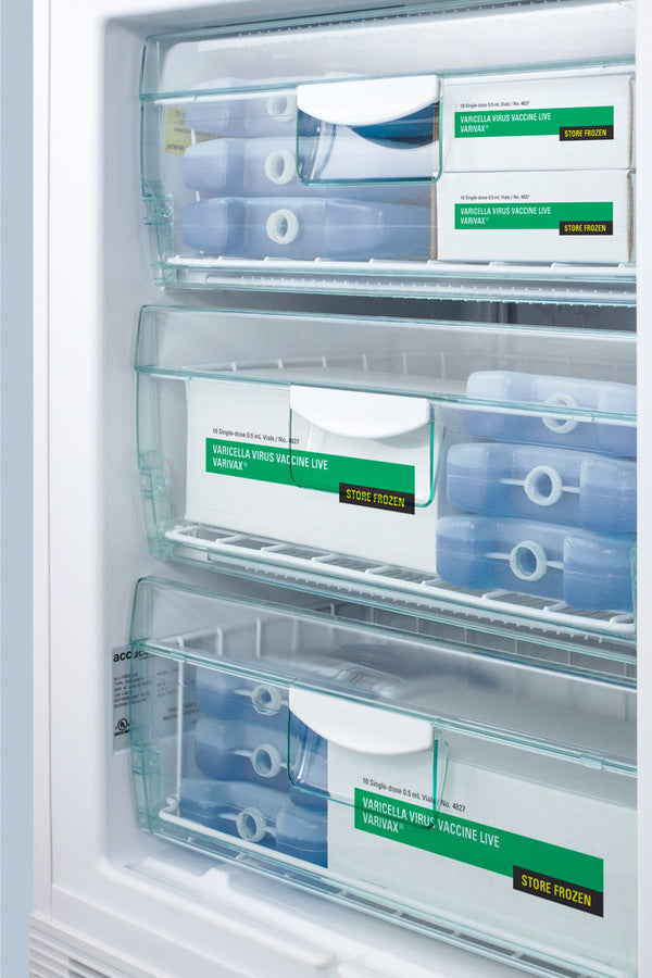 Three transparent freezer drawers contain boxes labeled Varicella Virus Vaccine Live and Store Frozen, along with several blue cold packs. The drawers are organized, and the vaccine boxes are white with green and yellow labels, kept neatly in an Accucold Medical Freezer. This undercounter all-freezer is ADA compliant, ensuring reliable storage within a medical setting.