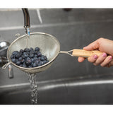 A hand holds a double fine mesh strainer with a wooden handle under running water in a sink, rinsing fresh blueberries. The water flows through the mesh, cleaning the berries. The person's fingernails are painted light pink.