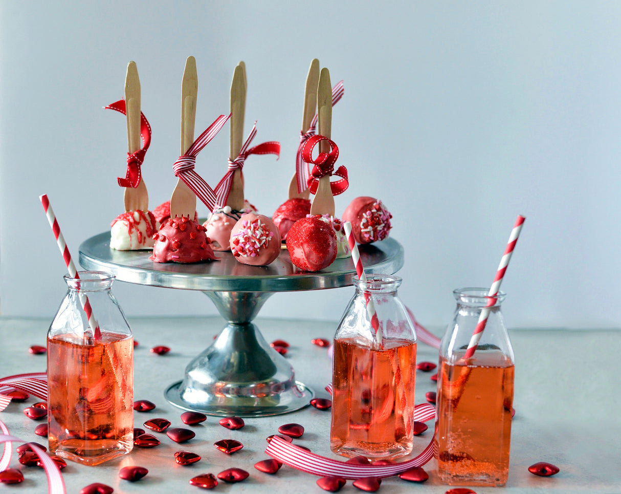 A stainless steel cake stand holds red and white cake pops tied with red ribbons and wooden forks. In front, glass bottles with pink drinks and red-striped straws sit among heart-shaped candies and striped ribbons on a light surface.