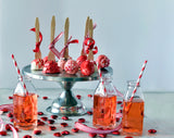 A stainless steel cake stand holds red and white cake pops tied with red ribbons and wooden forks. In front, glass bottles with pink drinks and red-striped straws sit among heart-shaped candies and striped ribbons on a light surface.