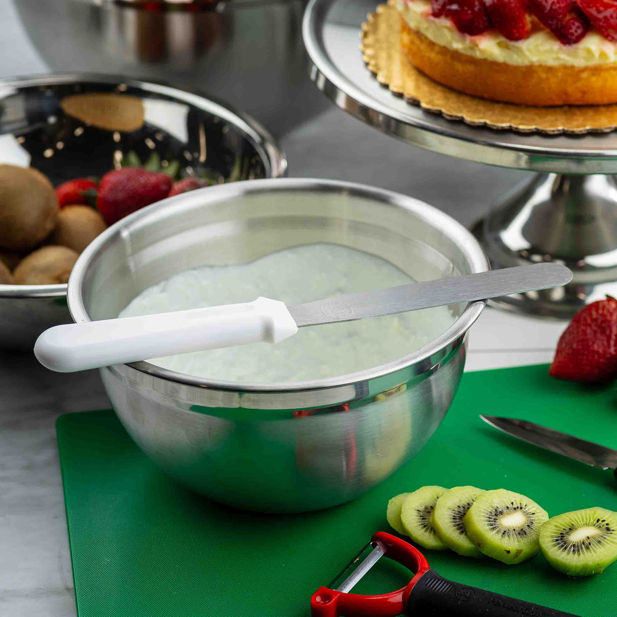 A 4 qt stainless steel mixing bowl filled with whipped frosting sits on a counter. In front are sliced kiwis, a peeler, and a knife on a green cutting board. Behind is a fruit tart and another metal bowl with whole kiwis and strawberries.