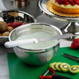 A 4 qt stainless steel mixing bowl filled with whipped frosting sits on a counter. In front are sliced kiwis, a peeler, and a knife on a green cutting board. Behind is a fruit tart and another metal bowl with whole kiwis and strawberries.