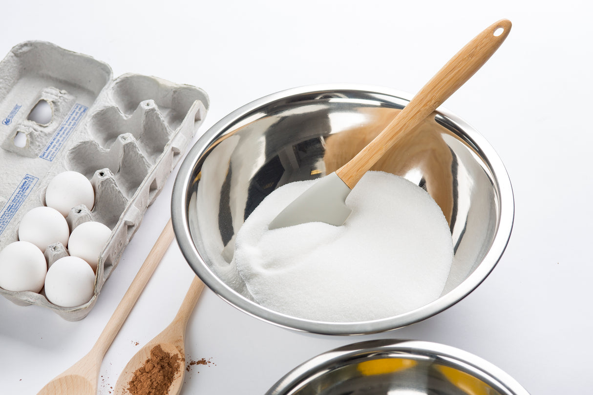 A stainless steel mixing bowl filled with white sugar and a spatula sits beside a carton of four white eggs, a wooden spoon with cocoa powder, and part of another bowl, all arranged on a white surface.