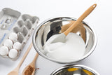 A stainless steel mixing bowl filled with white sugar and a spatula sits beside a carton of four white eggs, a wooden spoon with cocoa powder, and part of another bowl, all arranged on a white surface.