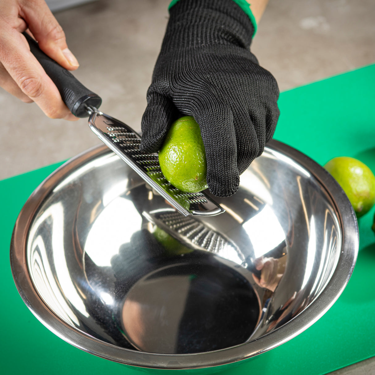 A person wearing black cut-resistant gloves is holding a lime and grating it over a stainless steel Mixing Bowl. The grater rests on the bowl’s edge, and a green cutting board with a whole lime sits in the background.