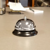 A shiny TableCraft chrome plated call bell with a black base sits on a light gray countertop. In the blurry background, a notebook, pen, and office items can be seen. The focus is mainly on the bell in the foreground.