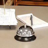 A chrome plated call bell sits on a countertop. Behind it are two restaurant guest checks, one on a stand and another flat with a pen on top. The TableCraft bell and papers rest on a smooth gray surface in a reception or cashier area.