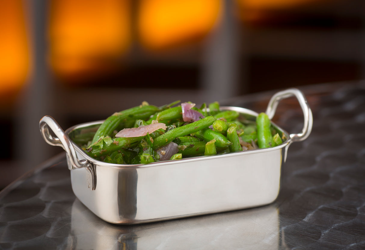 A shiny rectangular stainless steel mini roast pan with handles holds sautéed green beans and sliced red onions, placed on a reflective metal surface. The background is softly blurred with warm orange and brown tones.