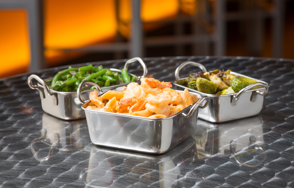 Three small stainless steel dishes, like mini roast pans, sit on a reflective table with sautéed green beans, grilled zucchini, and shrimp pasta. The softly lit background glows with warm orange tones, gently blurred for a cozy ambiance.