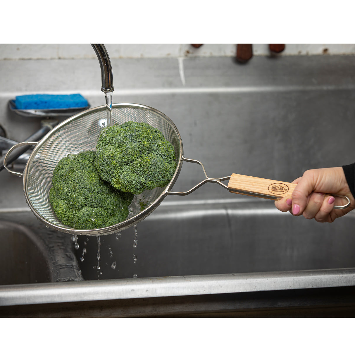 A hand holds a metal strainer with a wooden handle and medium mesh, rinsing two broccoli crowns under a running faucet above a stainless steel sink. Water droplets fall as a blue sponge rests on the sink’s ledge in the background.