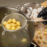 A hand in a black glove holds a fine mesh strainer with cooked gnocchi over a pot of hot water. In the background, eggs and uncooked gnocchi rest on a floured wooden surface as steam rises, highlighting the active cooking process.