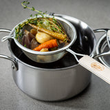 A fine mesh strainer with a wooden handle holds cooked carrots, onions, herbs, and mushrooms over a stainless steel pot filled with dark broth. The scene is set on a smooth gray countertop.