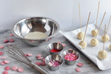 A marble surface holds a whisk, pink candy melts, a metal bowl with white chocolate, bowls of colorful sprinkles, and uncoated cake pops on 6 inch skewers resting on a lined tray, all ready for decorating. The background is bright and minimal.