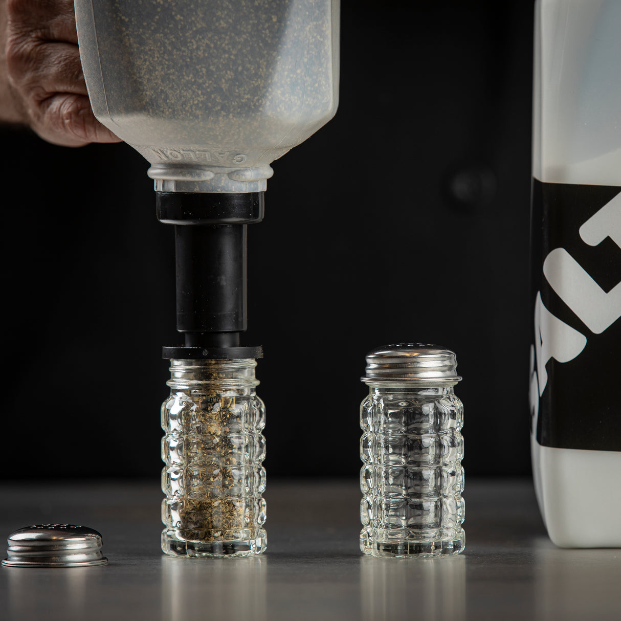 A hand uses a large pepper refiller to fill a clear, ridged glass spice jar with seasoning. Another empty, dishwasher safe spice jar with a metal lid rests nearby on a gray surface. A white container labeled SALT is partially visible on the right.