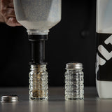 A hand uses a large pepper refiller to fill a clear, ridged glass spice jar with seasoning. Another empty, dishwasher safe spice jar with a metal lid rests nearby on a gray surface. A white container labeled SALT is partially visible on the right.
