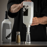 A person in a black shirt refills a transparent pepper shaker, made in USA, using a large plastic dispenser labeled PEP. Another dispenser and the shaker cap are on the table. The image focuses on hands as pepper pours into the shaker.