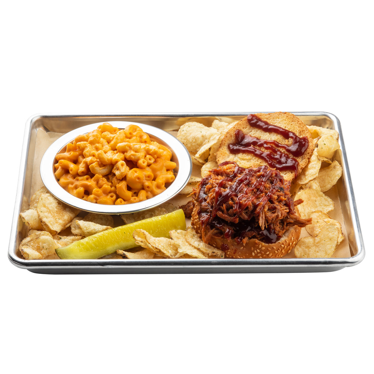 A 1/4 size aluminum sheet pan holds a pulled pork sandwich with barbecue sauce on a sesame bun, a pile of potato chips, creamy macaroni and cheese in a small bowl, and a pickle spear, all arranged neatly on parchment paper.