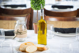 A Prima Bottle of olive oil with a stainless steel pourer sits on a white tablecloth beside sliced bread, a mason jar wrapped with twine, empty glasses, and a small potted plant; chairs are in the blurred background.