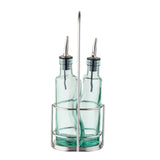 Two green tinted glass bottles with stainless steel pourers sit side by side in a silver metal caddy with a tall handle. The bottles appear empty and are likely intended for olive oil or vinegar. The design is simple and modern.