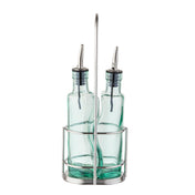 Two green tinted glass bottles with stainless steel pourers sit side by side in a silver metal caddy with a tall handle. The bottles appear empty and are likely intended for olive oil or vinegar. The design is simple and modern.