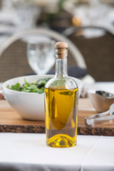 A clear glass bottle filled with golden olive oil and sealed with a cork stopper stands on a white tablecloth. Behind it, a white bowl of salad greens, metal salad tongs, and a drinking glass from TableCraft Products H9220 sit slightly out of focus.