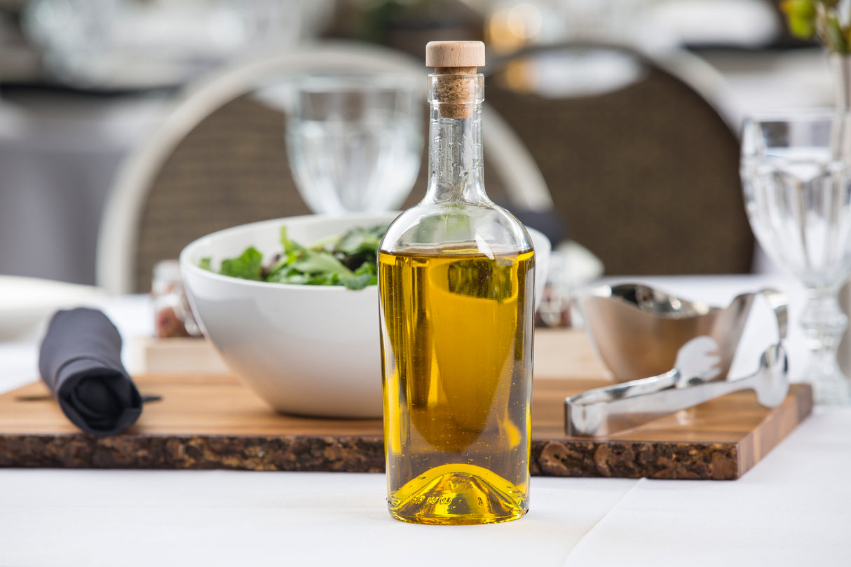 A clear TableCraft glass bottle filled with golden olive oil, sealed with a cork stopper, sits on a dining table. In the background, a white bowl of leafy green salad, utensils, and glassware suggest an inviting meal setting.