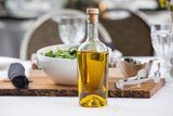 A clear TableCraft glass bottle filled with golden olive oil, sealed with a cork stopper, sits on a dining table. In the background, a white bowl of leafy green salad, utensils, and glassware suggest an inviting meal setting.