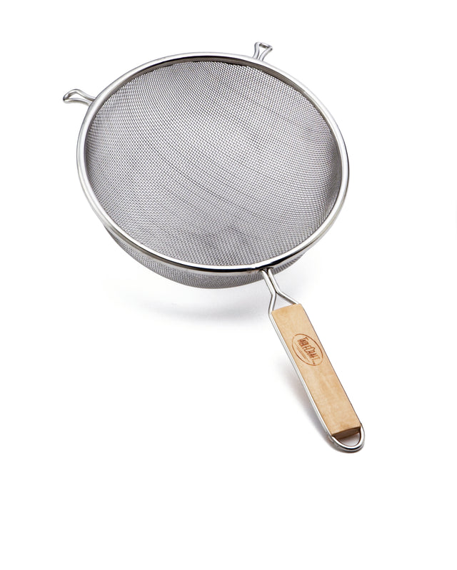 A stainless steel strainer with a wooden handle and two metal hooks on the rim is shown on a white background. The handle features a logo, and the strainer is angled to display its fine mesh and sturdy construction.