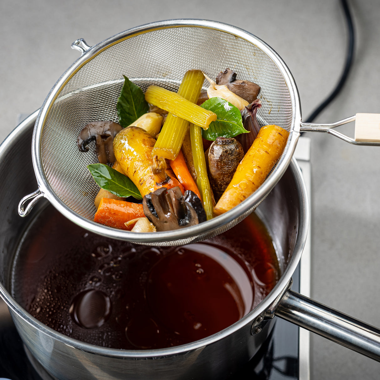 A fine mesh strainer with a wooden handle holds cooked vegetables—carrots, celery, mushrooms, and herbs—over a pot of dark broth. The vegetables are being drained, showing homemade stock in progress. The kitchen surface and pot handles are visible.
