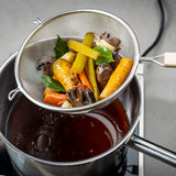 A fine mesh strainer with a wooden handle holds cooked vegetables—carrots, celery, mushrooms, and herbs—over a pot of dark broth. The vegetables are being drained, showing homemade stock in progress. The kitchen surface and pot handles are visible.