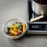 A fine mesh strainer with a light wooden handle holds cooked carrots, celery, mushrooms, onions, and bay leaves on a gray counter beside an induction cooktop displaying “0.0.0” with a pot of brown liquid.