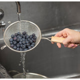 A hand holds a double medium mesh strainer with a wooden handle under a running faucet, rinsing fresh blueberries in the kitchen sink. Water streams through the mesh, thoroughly washing the berries as the sink and faucet appear in the background.