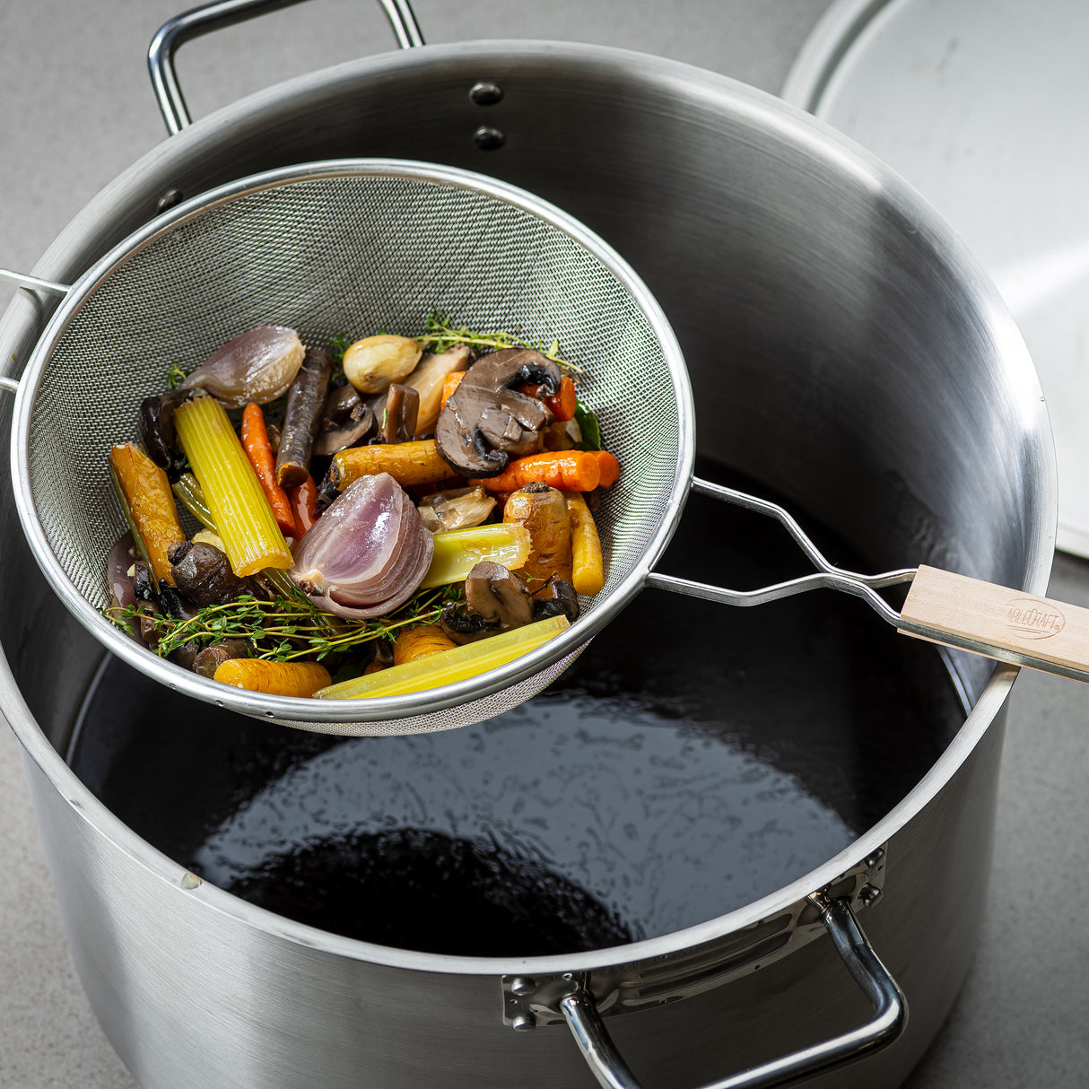 A fine mesh strainer with a wooden handle holds cooked vegetables—carrots, celery, onions, garlic, and mushrooms—above a large stainless steel pot of dark broth. The pot sits on a gray countertop, its lid resting to one side.