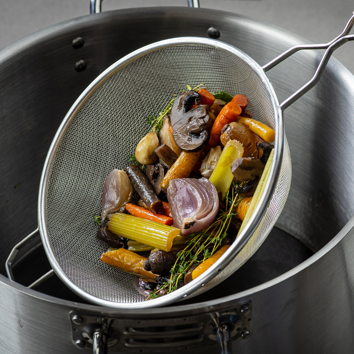 A fine mesh strainer with a wooden handle, filled with roasted carrots, celery, mushrooms, shallots, and herbs, rests over a large stainless steel pot—perfect for making rich vegetable broth. The vegetables appear cooked and slightly browned.