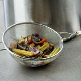 A metal strainer with a fine mesh and wooden handle, filled with cooked carrots, celery, onions, mushrooms, and herbs, rests on a gray countertop in front of a large stainless steel pot. The vegetables appear soft after straining.