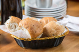 A hand-woven black basket with gold trim, lined with white napkins, holds four golden-brown bread rolls on a wooden table. In the blurred background, stacked white plates and a silver cup suggest a table set for a meal.