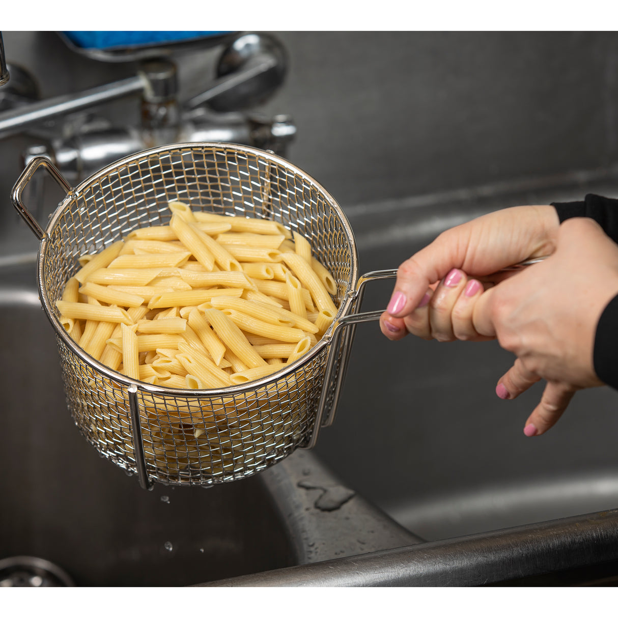 A person holds a stainless steel cooking basket filled with cooked penne pasta over the sink, draining water. Their hands grip the handles as droplets fall into the stainless steel sink below. The basket is also dishwasher safe for easy cleaning.