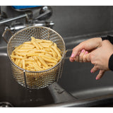 A person holds a stainless steel cooking basket filled with cooked penne pasta over the sink, draining water. Their hands grip the handles as droplets fall into the stainless steel sink below. The basket is also dishwasher safe for easy cleaning.